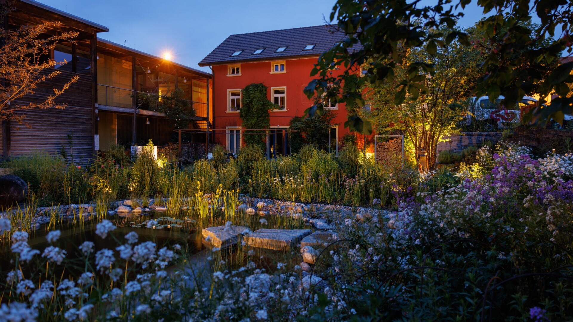 Ein Garten mit einem von Blumen und Grün umgebenen Teich wird in der Abenddämmerung beleuchtet, mit einem roten Haus und einem Holzgebäude im Hintergrund unter einem blauen Abendhimmel.