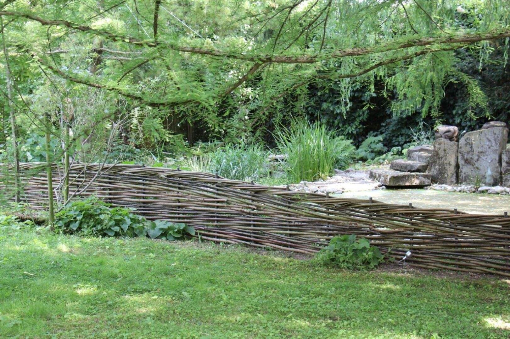 A rustic, woven wooden fence borders a lush garden with green grass, leafy plants and tall trees. In the background are stones and dense greenery that create a calm, natural atmosphere.
