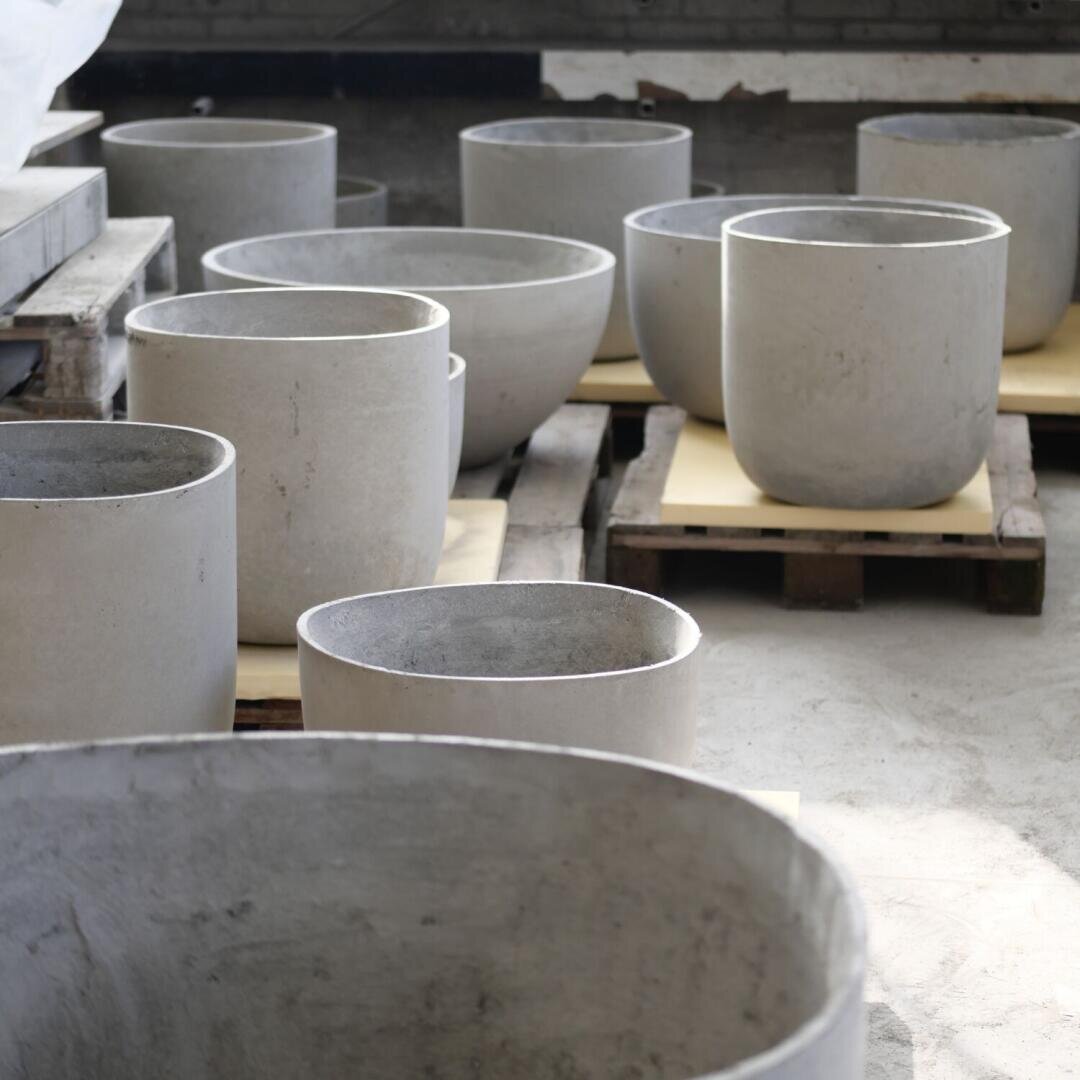Große, runde, hellgraue Betonpflanzgefäße in verschiedenen Größen sind auf Holzpaletten in einem hellen Innenraum mit Betonboden angeordnet.