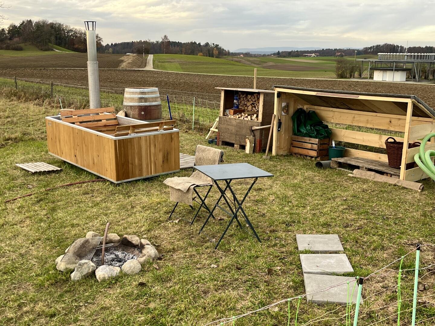 The rustic outdoor area consists of a wooden hot tub, a small table with two chairs, a fireplace surrounded by stones, a wood storage shed and garden tools on a meadow with fields in the background.