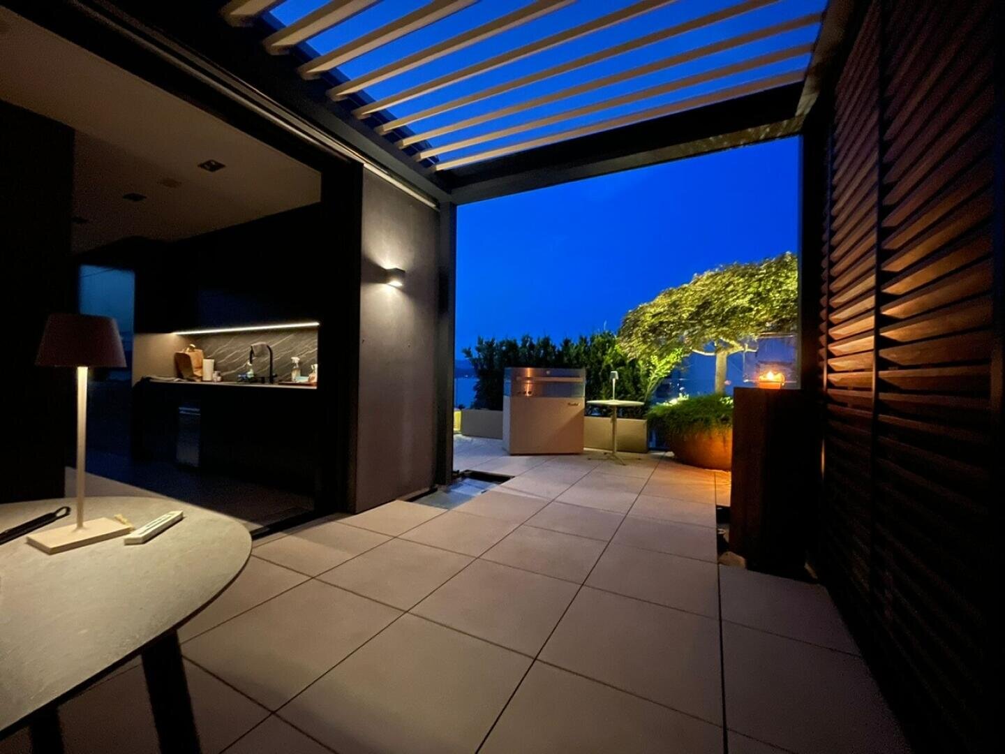 Eine moderne Außenterrasse in der Abenddämmerung mit gefliestem Boden, einer teilweise überdachten Pergola, einem Küchenbereich, Topfpflanzen, warmer Beleuchtung und einem Blick in den Abendhimmel. Auf der linken Seite ist ein gemütlicher Innenbereich zu sehen.