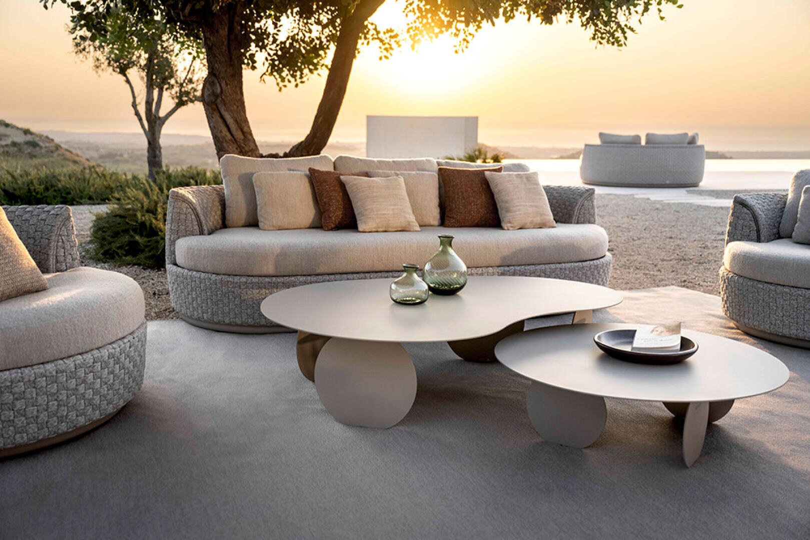 Moderner Lounge-Bereich im Freien mit geflochtenen Sofas, neutralen Kissen und zwei runden Couchtischen mit dekorativen Vasen auf einer Terrasse mit Bäumen und einem malerischen Blick auf den Sonnenuntergang im Hintergrund.