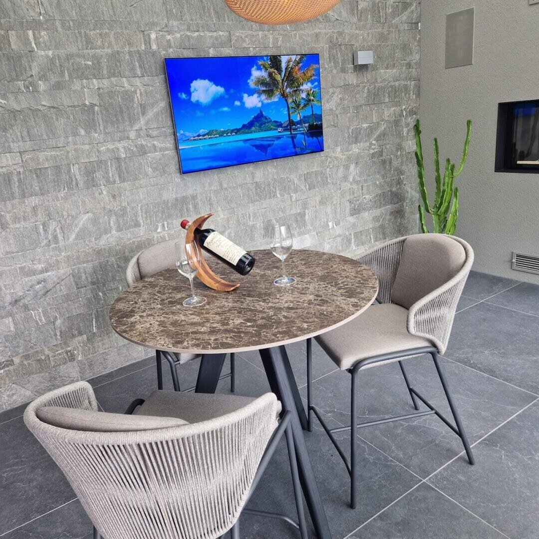 Ein runder Marmortisch mit zwei Stühlen, einer Weinflasche in einem Halter und zwei Weingläsern steht auf einem grauen Kachelboden. Ein Fernseher an der Steinwand zeigt eine tropische Strandszene. In der Ecke steht ein großer Kaktus.