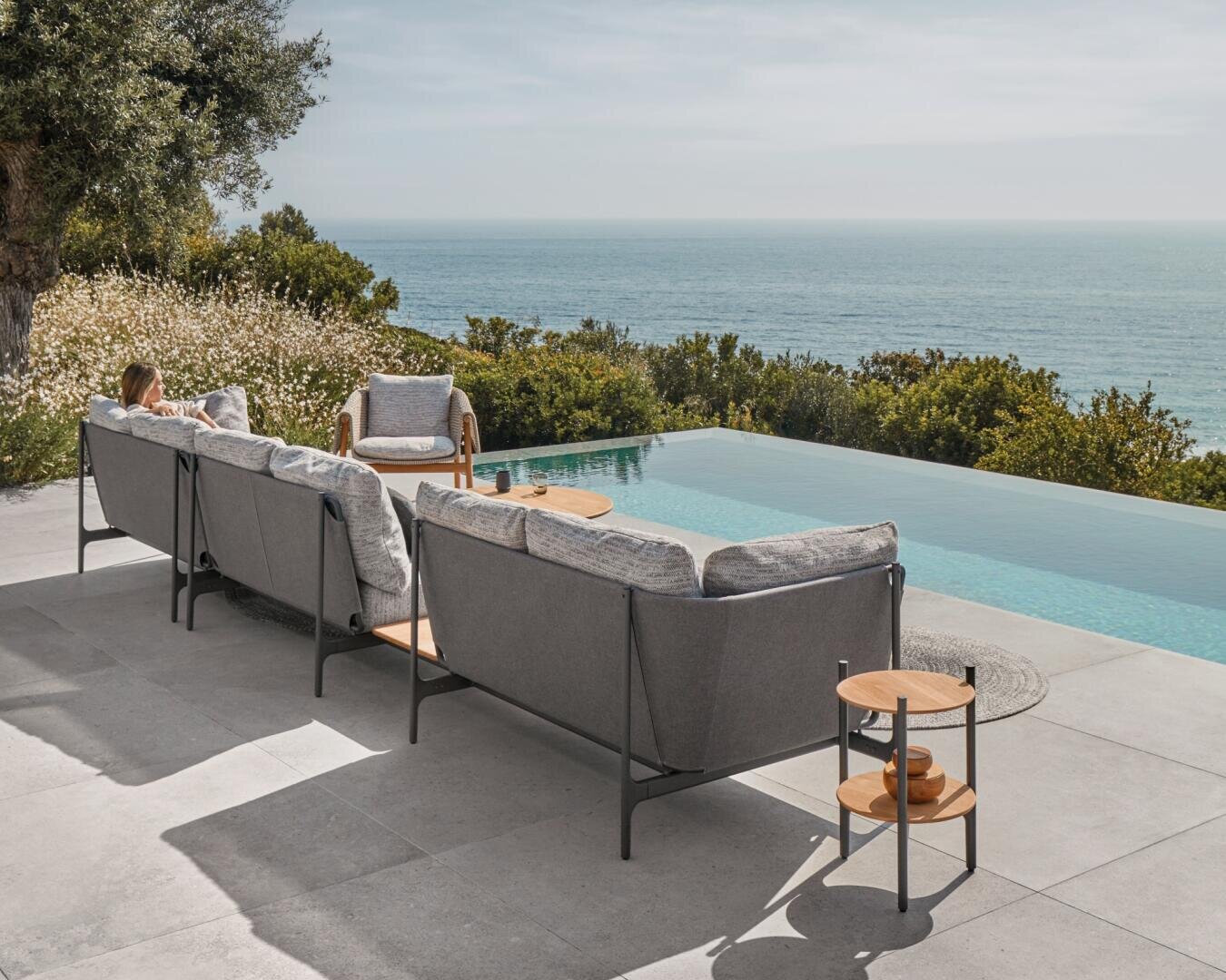 Eine Person entspannt sich an einem sonnigen Tag auf einem modernen Außensofa an einem Infinity-Pool mit Blick auf den Ozean und das Grün der Bäume im Hintergrund.