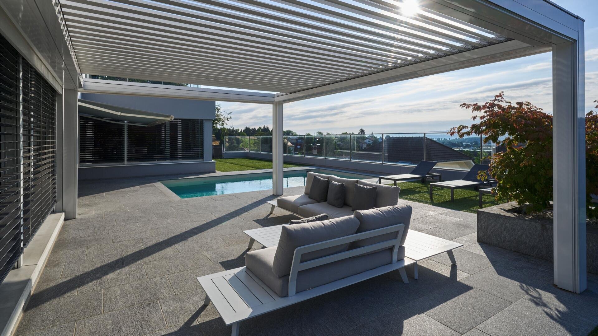 Modern outdoor patio with white pergola, cushioned lounge seating, and a pool overlooking rooftops and distant views, under a partly cloudy sky. Sunlight casts shadows across the stone-tiled floor.