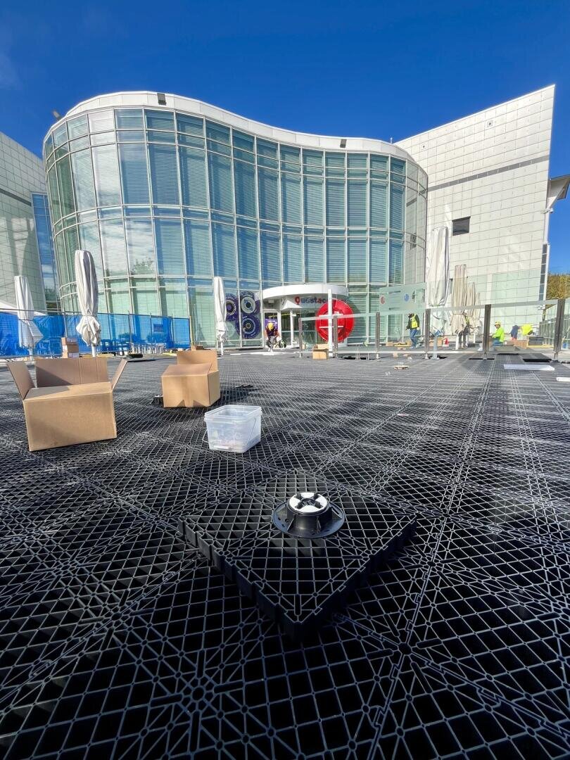 Modern glass building with curved façade, outdoor area with lattice flooring, brown chairs, closed parasols and scattered objects under a clear blue sky.