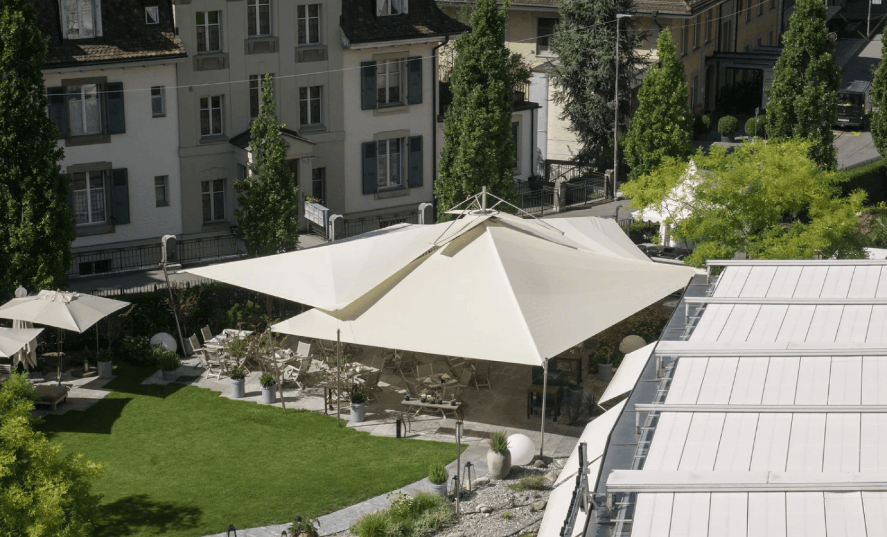 A large, white, modern canopy shades an outdoor seating area in a green garden, surrounded by buildings and tall trees on a sunny day.