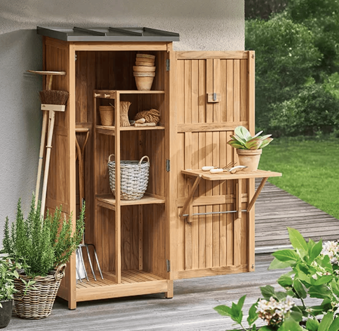 Ein hölzerner Aufbewahrungsschrank für den Außenbereich mit geöffneter Tür, in der sich Regale mit Gartengeräten, Körben und Töpfen befinden. Auf einem kleinen ausklappbaren Tisch an der Tür steht eine Topfpflanze. Üppiges Grün umgibt den Bereich.