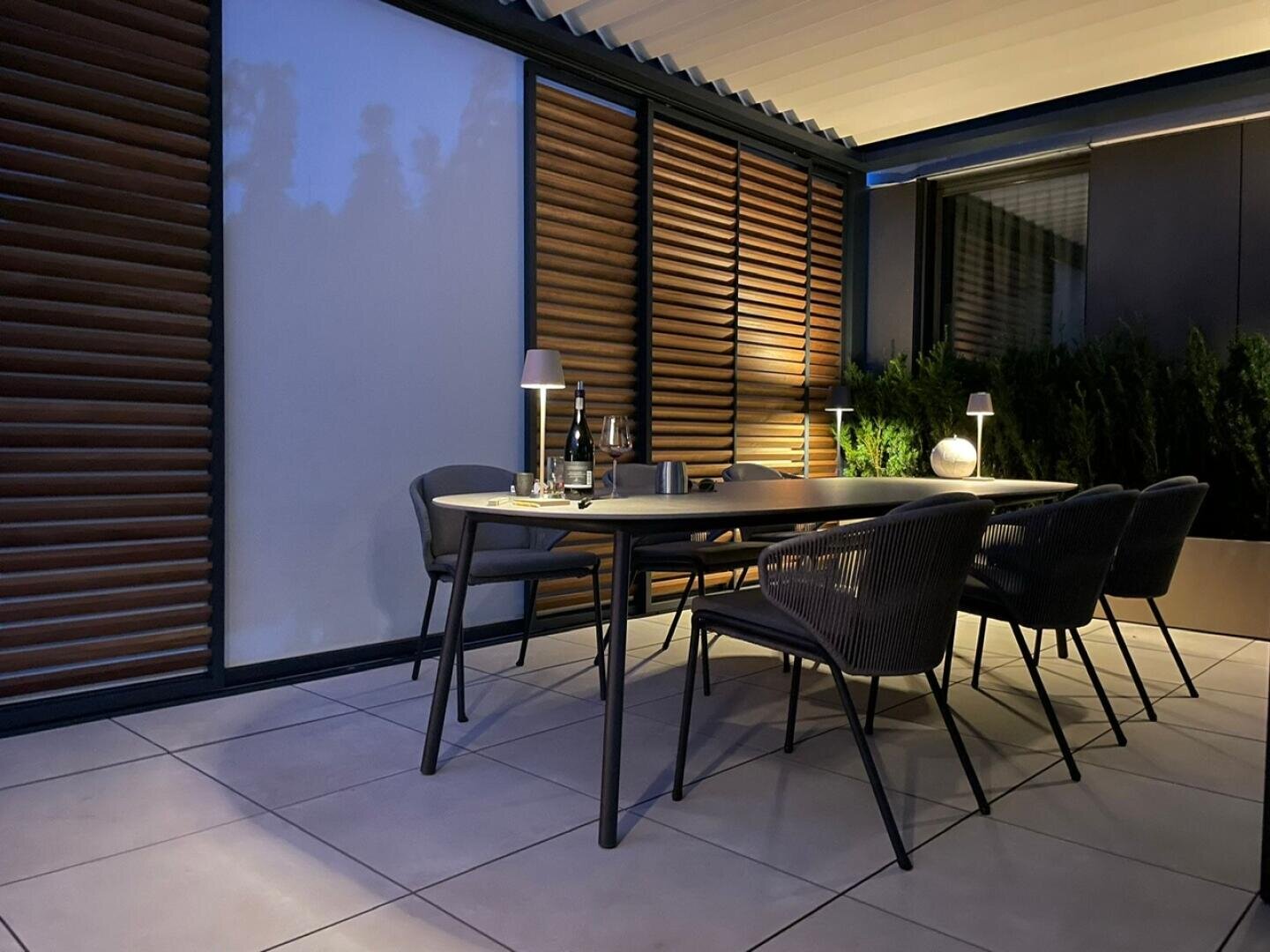 Ein moderner Essbereich im Freien in der Abenddämmerung mit einem rechteckigen Tisch, sechs Stühlen, Weingläsern, Flaschen und kleinen Lampen. Der Raum ist von Glaswänden und hölzernen Fensterläden umgeben, mit sanfter Beleuchtung und Topfpflanzen im Hintergrund.
