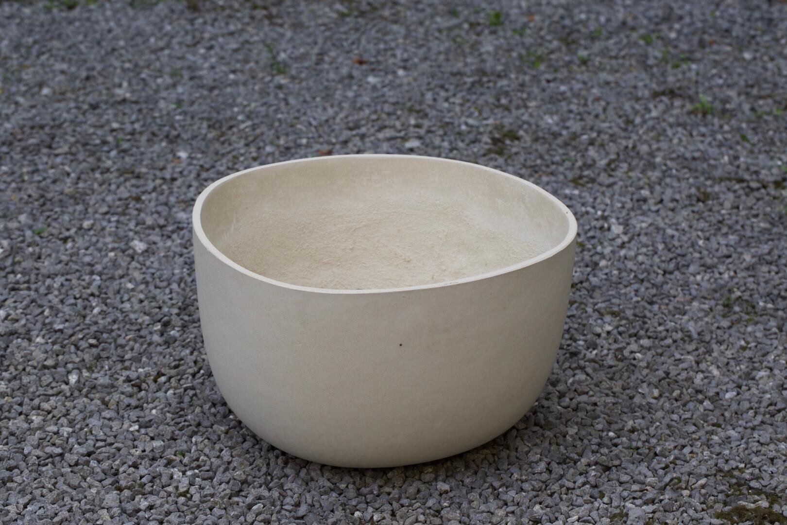 Eine große, runde, cremefarbene Keramikschale steht leer auf einer mit kleinen grauen Kieselsteinen bedeckten Fläche.