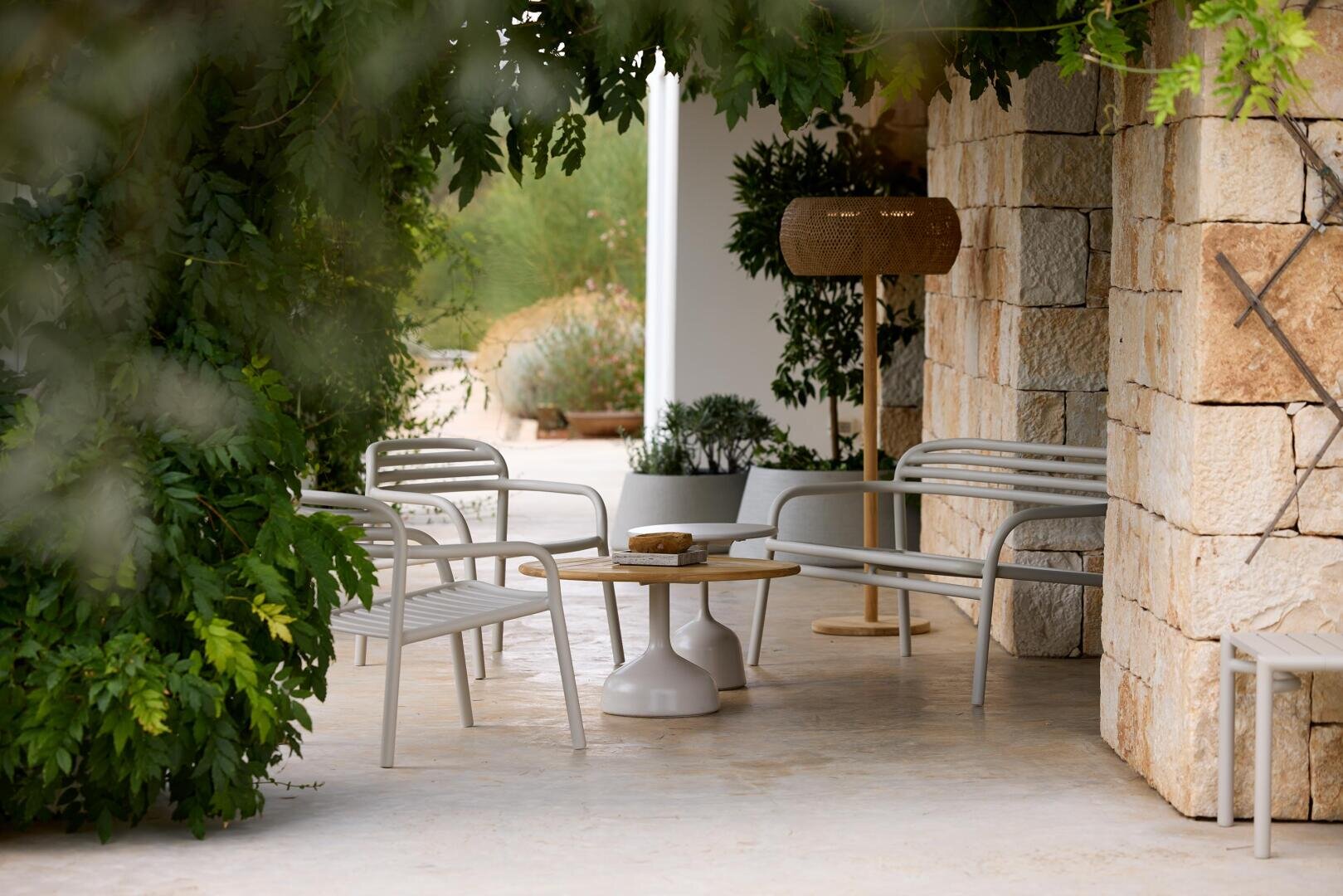 Eine schattige Terrasse mit Metallbänken, einem runden Tisch, Topfpflanzen und einer Stehlampe aus Korbgeflecht, umgeben von Steinmauern und üppigem Grün.