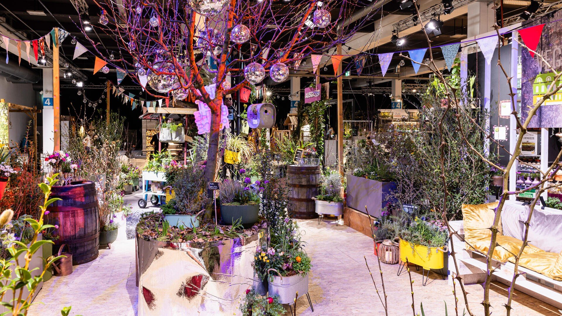 Eine lebhafte Indoor-Gartenszene mit Topfpflanzen, Bäumen und Blumen, bunten Wimpeln, hängenden Discokugeln, Holzfässern und einer gemütlichen gelben Sitzecke, alles unter heller dekorativer Beleuchtung.