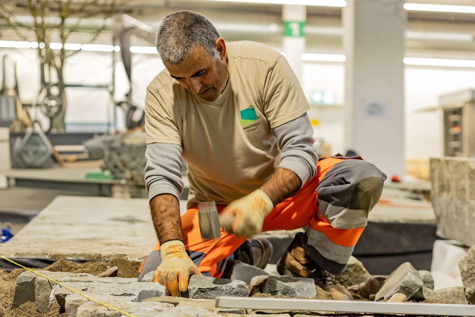 Ein Bauarbeiter in orange-beiger Uniform kniet auf dem Boden und bearbeitet mit Hammer und Meißel graue Steine in einem Gebäude. Um ihn herum sind Werkzeuge und Baumaterialien zu sehen.