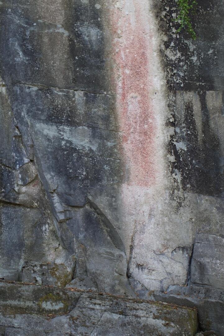 Eine vertikale Felswand mit verschiedenen Grau- und Brauntönen, die in der Mitte einen auffälligen vertikalen Streifen mit rosa-roter Färbung aufweist, umgeben von rauen, ungleichmäßigen Steinstrukturen.