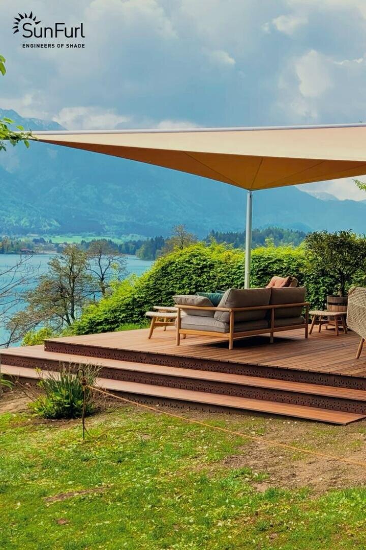 Eine Holzterrasse mit Sitzgelegenheiten im Freien wird von einem großen dreieckigen Vordach beschattet. Von der Terrasse aus hat man Blick auf einen malerischen See und die Berge, umgeben von üppigem Grün. Das SunFurl-Logo ist in der oberen linken Ecke zu sehen.
