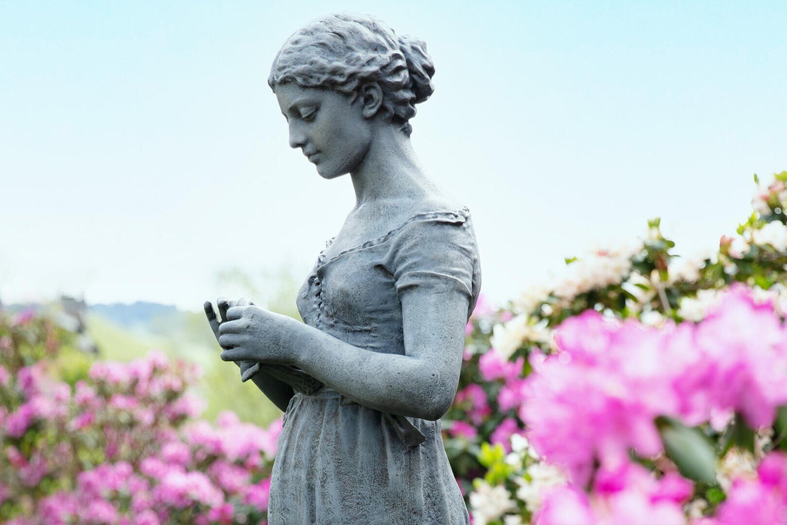 Eine steinerne Statue einer jungen Frau in einem einfachen Kleid steht inmitten blühender rosa Blumen und blickt mit friedlichem Gesichtsausdruck unter einem klaren Himmel auf ihre Hände herab.