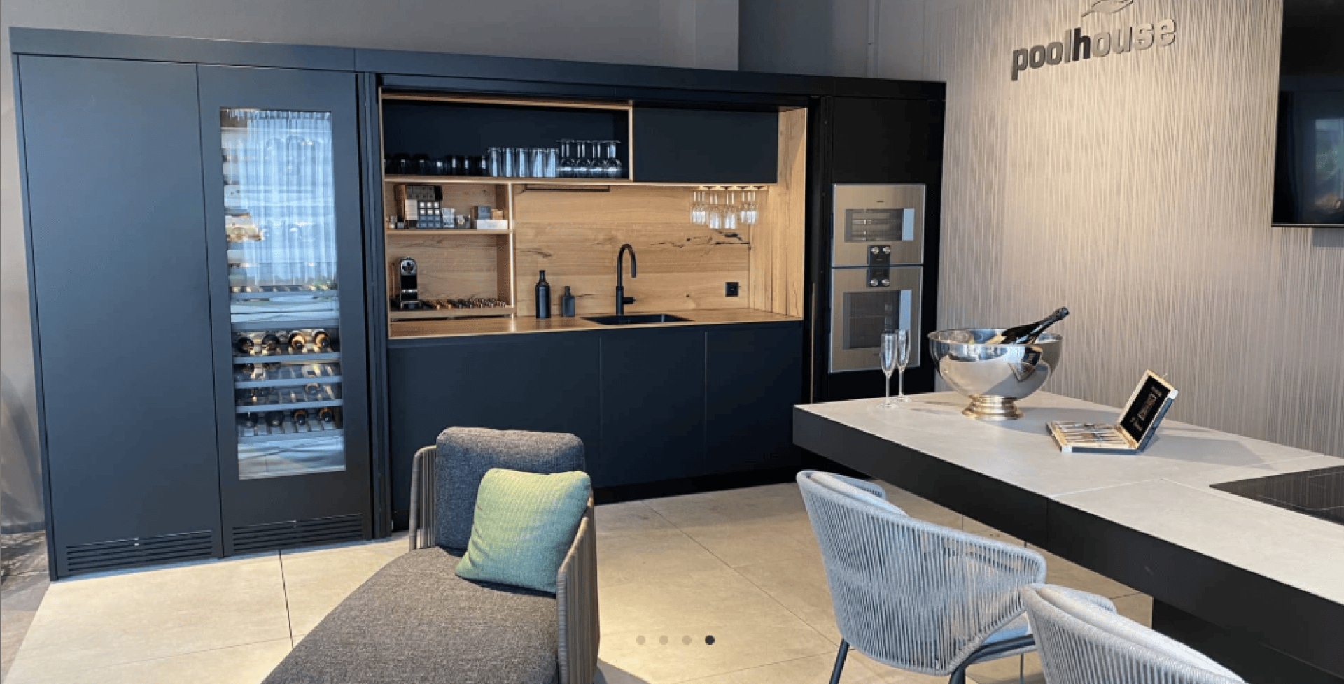 Moderne Küche mit dunklen Schränken, eingebautem Weinkühlschrank, Aufkantung aus Holz und offenen Regalen. Auf der Arbeitsplatte befinden sich ein Waschbecken und Glaswaren. In der Nähe sind eine graue Lounge-Sessel, Tisch mit Stühlen, und Wand-TV montiert.