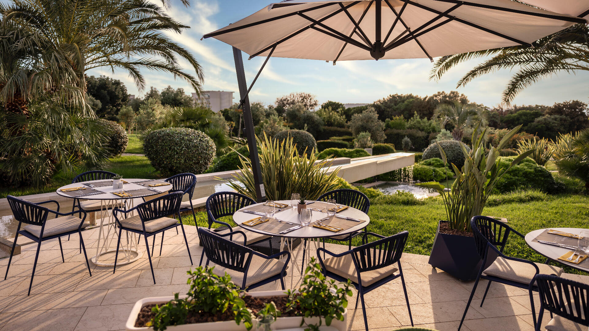 Restaurantterrasse mit runden Tischen und schwarzen Stühlen unter einem großen Sonnenschirm, umgeben von üppigem Grün, Palmen und gepflegten Sträuchern an einem sonnigen Tag.