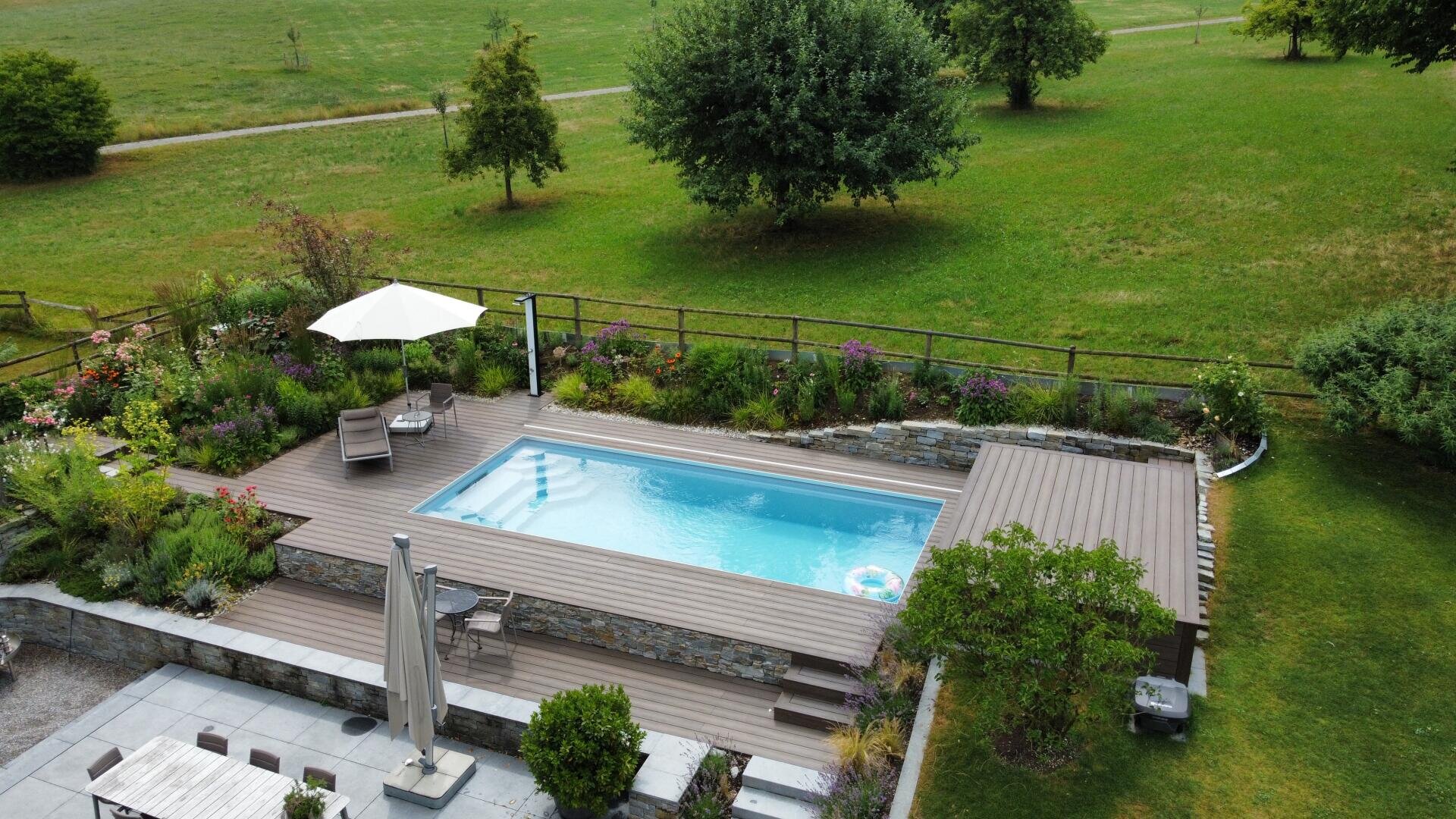 Ein kleiner rechteckiger Pool, umgeben von einer Holzterrasse, liegt in einem üppigen Garten mit grünem Gras, Bäumen, bunten Blumen, Liegestühlen und Sonnenschirmen in einer friedlichen Außenumgebung.