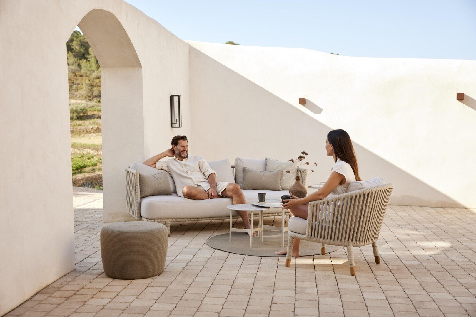 Ein Mann und eine Frau entspannen sich auf Terrassenmöbeln in einem sonnenbeschienenen Innenhof mit weißen Wänden und gewölbten Türöffnungen. Sie sitzen bequem und scheinen ein angenehmes Gespräch zu führen.