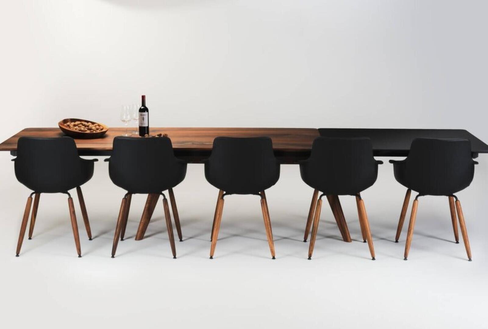 A modern dining table with a wooden top, six black chairs with wooden legs, a bowl of nuts, two wine glasses, and a bottle of wine arranged neatly on the table against a plain background.