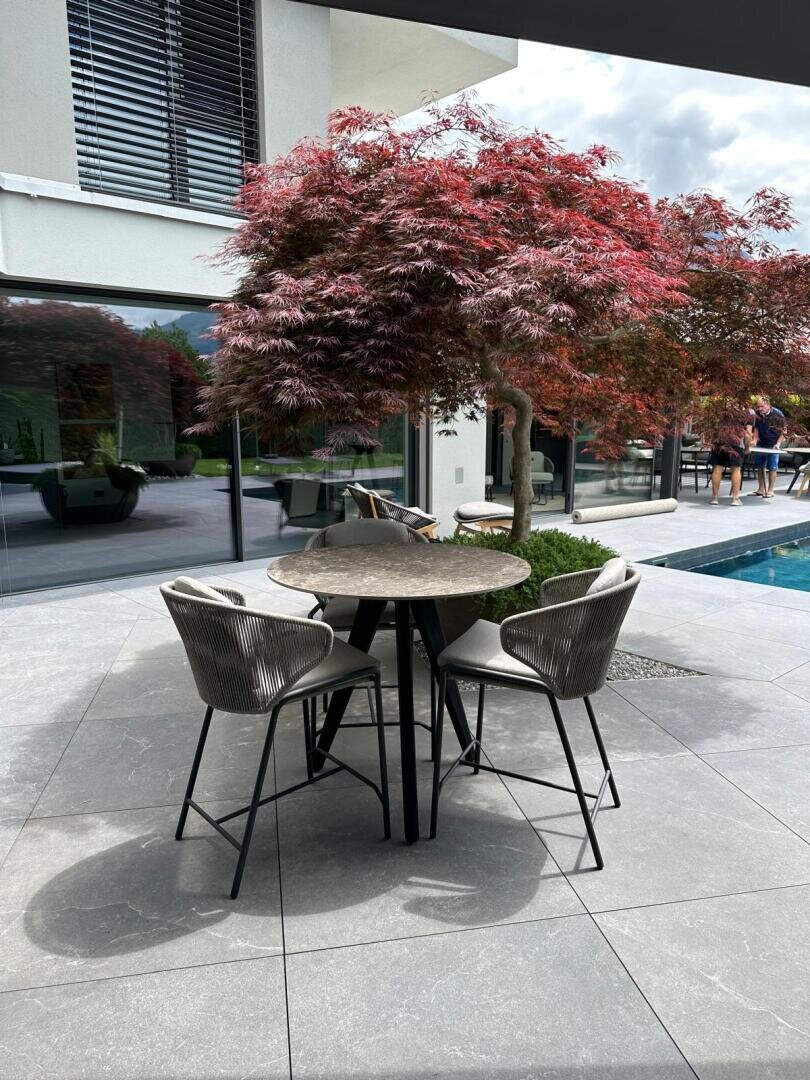 Eine moderne Außenterrasse mit einem runden Tisch und drei Stühlen steht auf einem gefliesten Boden neben einem Pool. Rotblättrige Bäume spenden Schatten, und die Terrasse grenzt an ein Gebäude mit Glaswänden.