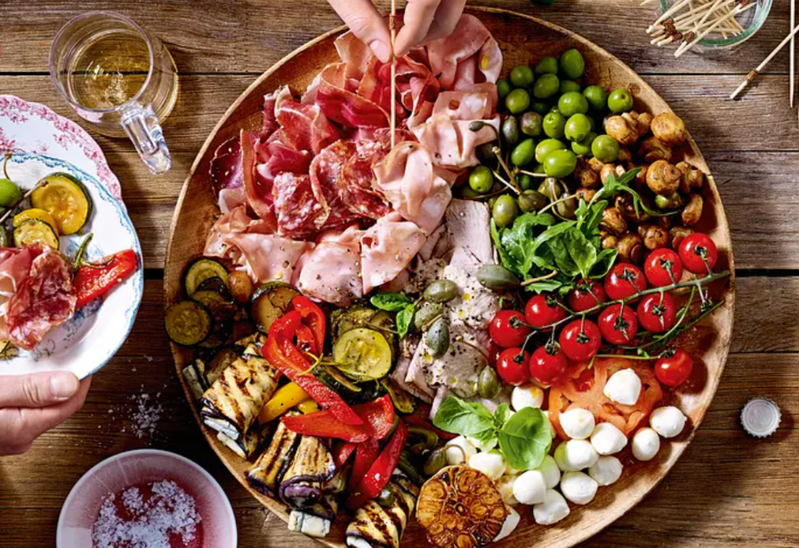 Eine Holzplatte, gefüllt mit Wurst, Käse, Kirschtomaten, Oliven, gegrilltem Gemüse und Kapern. Eine Hand serviert das Essen auf einem Teller, daneben stehen Getränke, Salz und Zahnstocher auf einem rustikalen Holztisch.