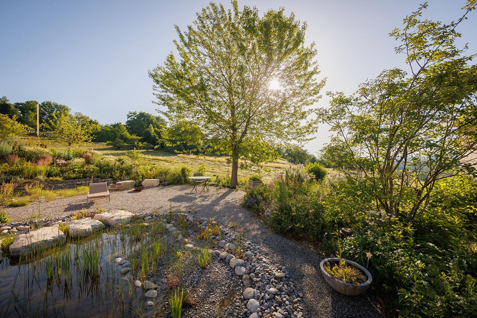 Ein sonnenbeschienener Garten mit einem großen Baum, Kieswegen, einem mit Steinen eingefassten Teich und verstreuten Stühlen; üppiges Grün und sanfte Hügel im Hintergrund unter einem klaren blauen Himmel.