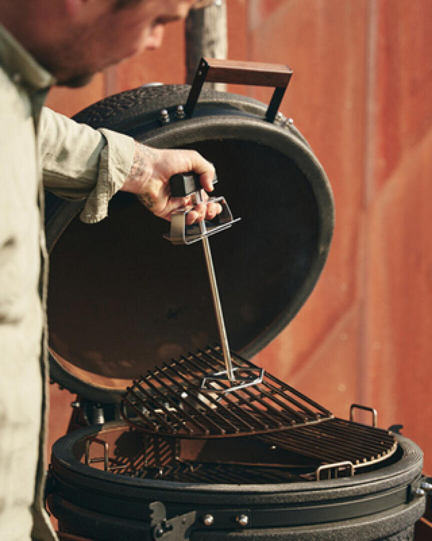 A person uses a metal tool to lift the grill grate of a round, black outdoor grill with an open lid against a blurred brown background.