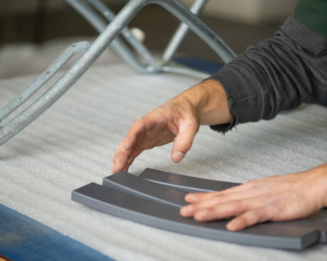 Eine Person arrangiert drei gebogene graue Metallteile auf einer gepolsterten Fläche, wobei ein Teil eines Metallrahmens und ihre Arme sichtbar sind.