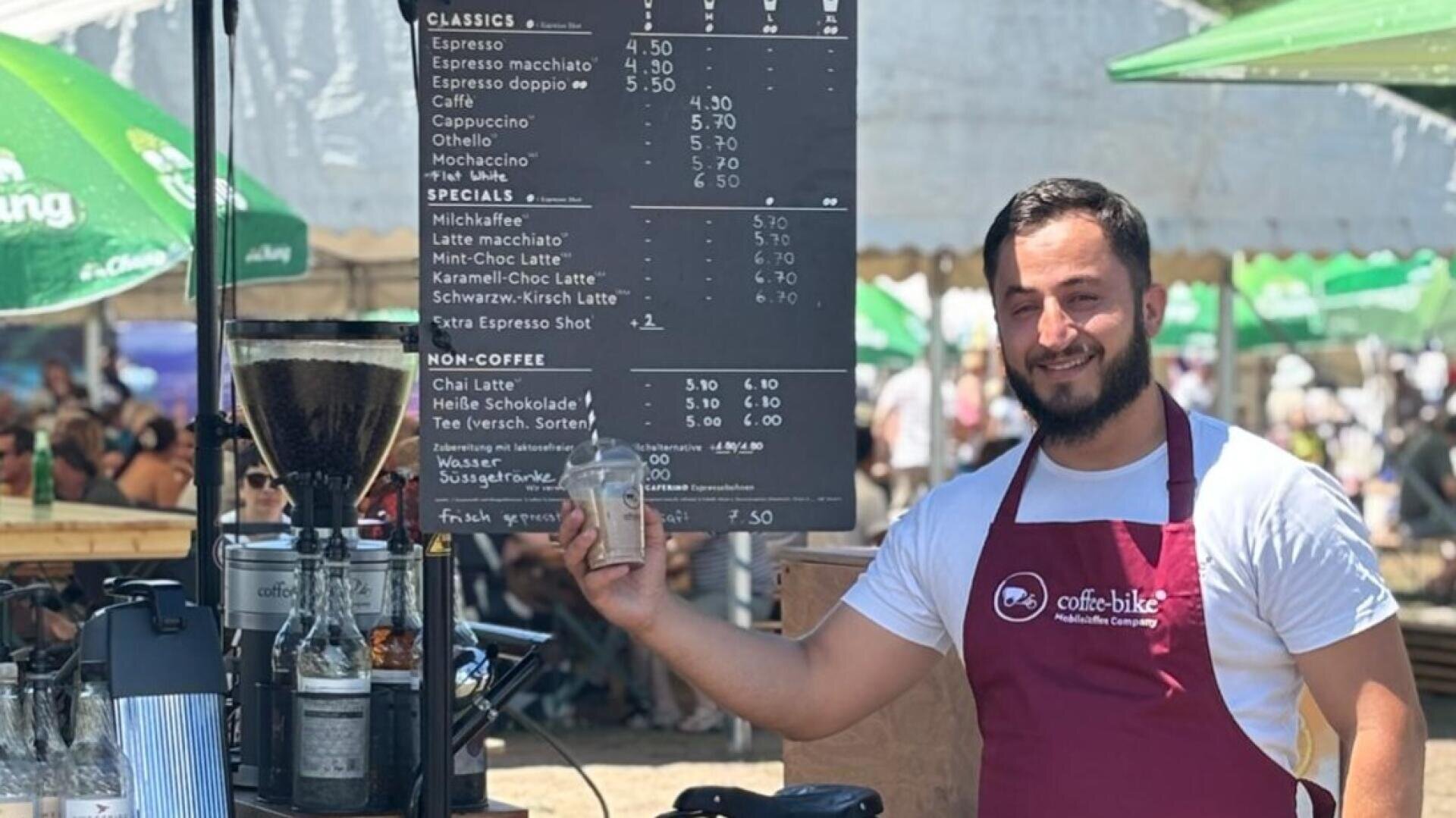 Ein lächelnder Barista in einer roten Schürze steht an einem Kaffeewagen im Freien und hält eine kleine Tasse in der Hand. Hinter ihm ist eine schwarze Menükarte mit Kaffeeoptionen zu sehen. Im Hintergrund der sonnigen Veranstaltung sind Menschen und grüne Regenschirme zu sehen.