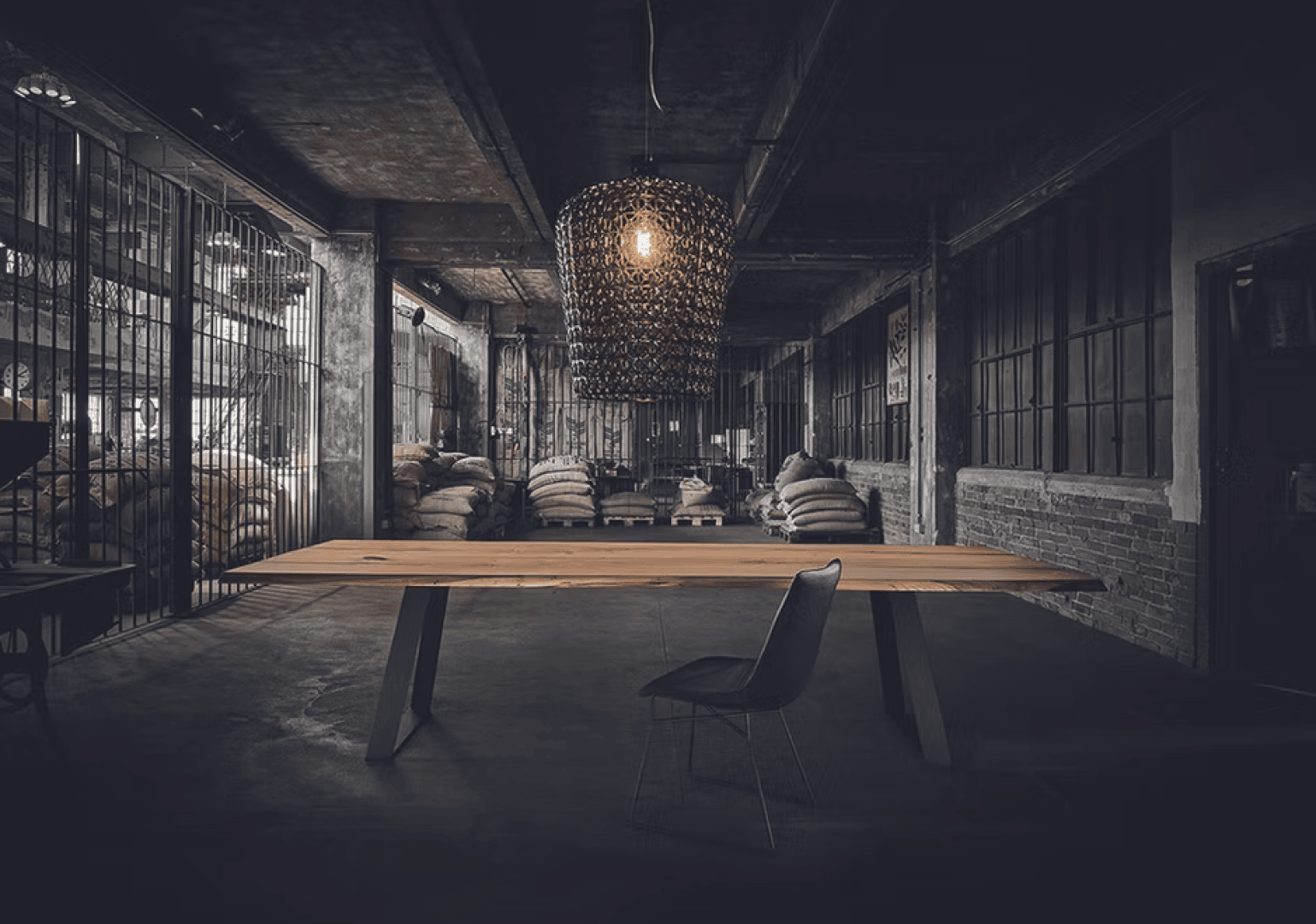 A single chair sits at a long wooden table in a dimly lit industrial warehouse with exposed brick, metal cages, and stacked burlap sacks in the background. A large, modern pendant light hangs above the table.