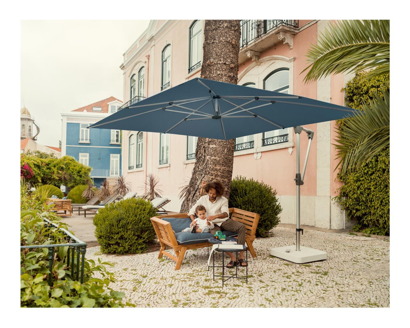 Eine Frau und ein Kind sitzen zusammen auf einer Holzbank unter einem großen blauen Sonnenschirm in einem sonnigen Innenhof mit pastellfarbenen Gebäuden, grünen Büschen und Palmen. In der Nähe steht ein kleiner Tisch mit Getränken.