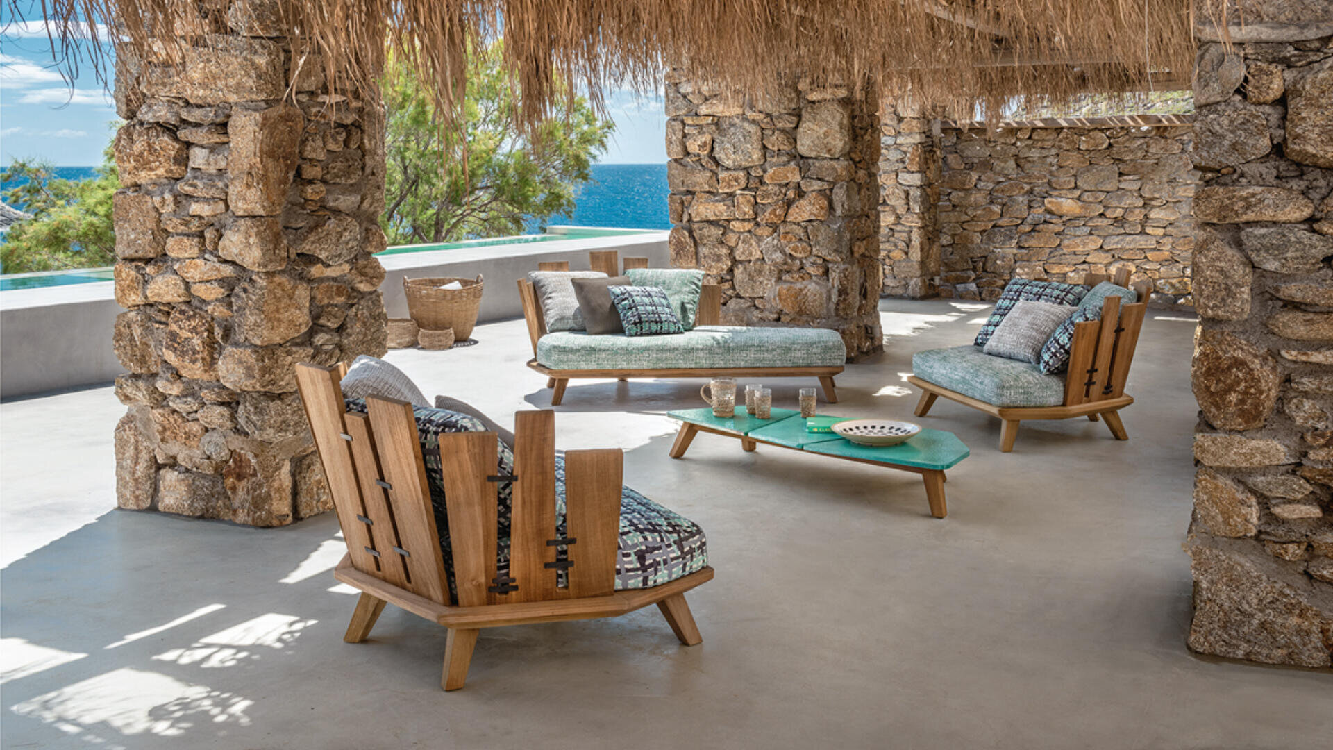 Außenbereich mit rustikalen Steinsäulen, Holzmöbeln mit gemusterten Kissen, einem niedrigen grünen Couchtisch und einem Strohdach, von dem aus man an einem sonnigen Tag das Meer sehen kann.