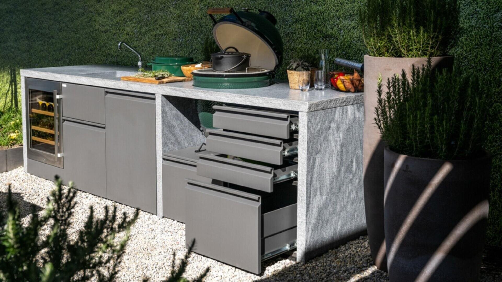 Modern outdoor kitchen with a marble countertop, open drawers, a small sink, a green ceramic grill, wine fridge, and potted plants, set against a green hedge background on gravel ground.