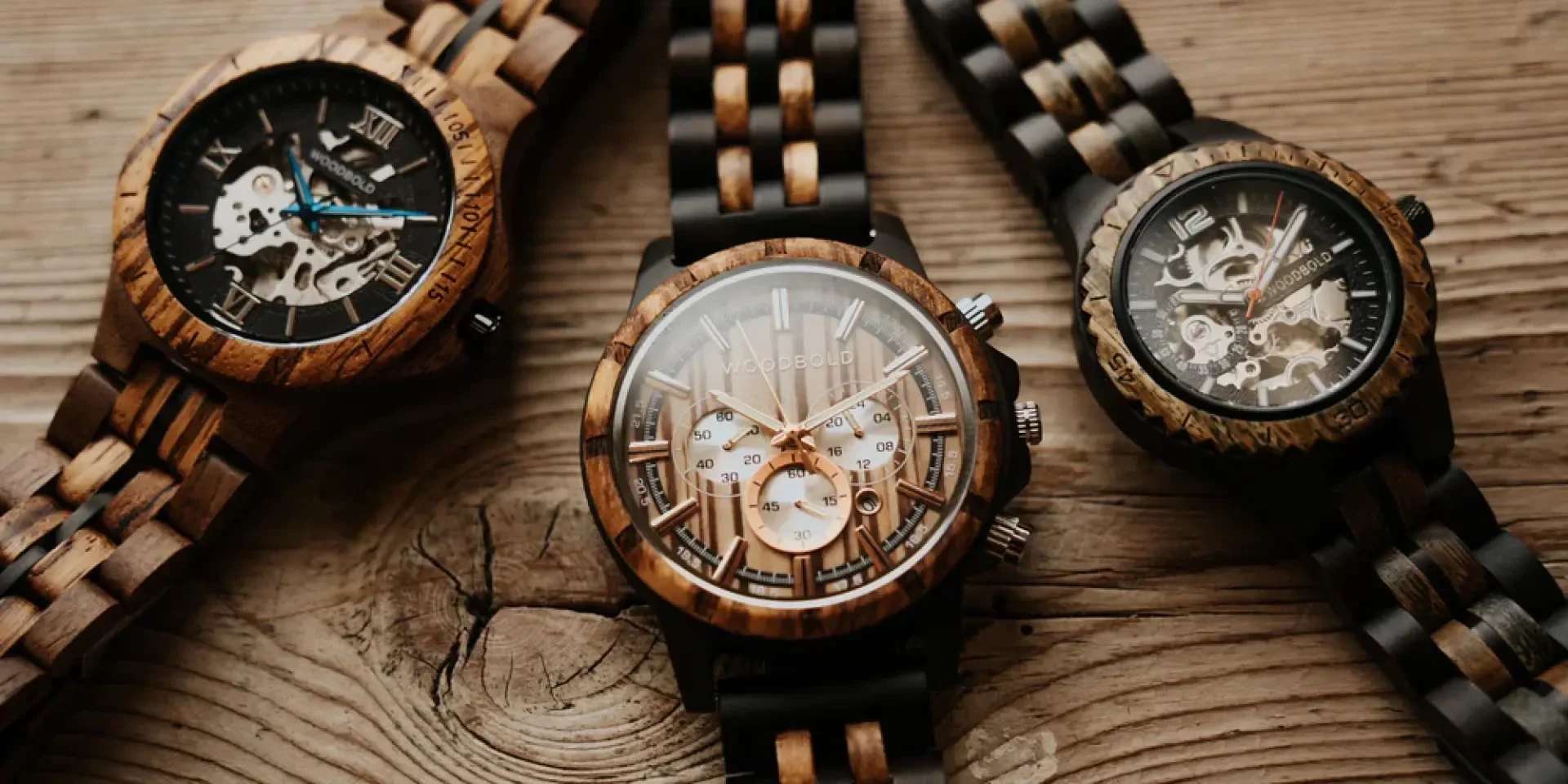 Three wooden wristwatches with exposed mechanical gears are displayed on a rustic wooden surface. The clocks feature a mixture of light and dark wooden straps and intricate open dials that reveal their inner workings.