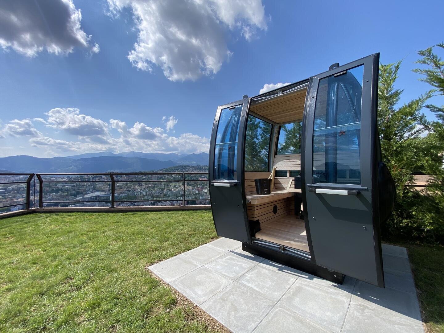 Eine moderne, schwarze Außensauna in Form einer Seilbahn steht auf einer Steinterrasse mit offener Tür und bietet einen Blick auf einen grasbewachsenen Hof, malerische Berge und eine Stadt unter einem blauen Himmel mit vereinzelten Wolken.