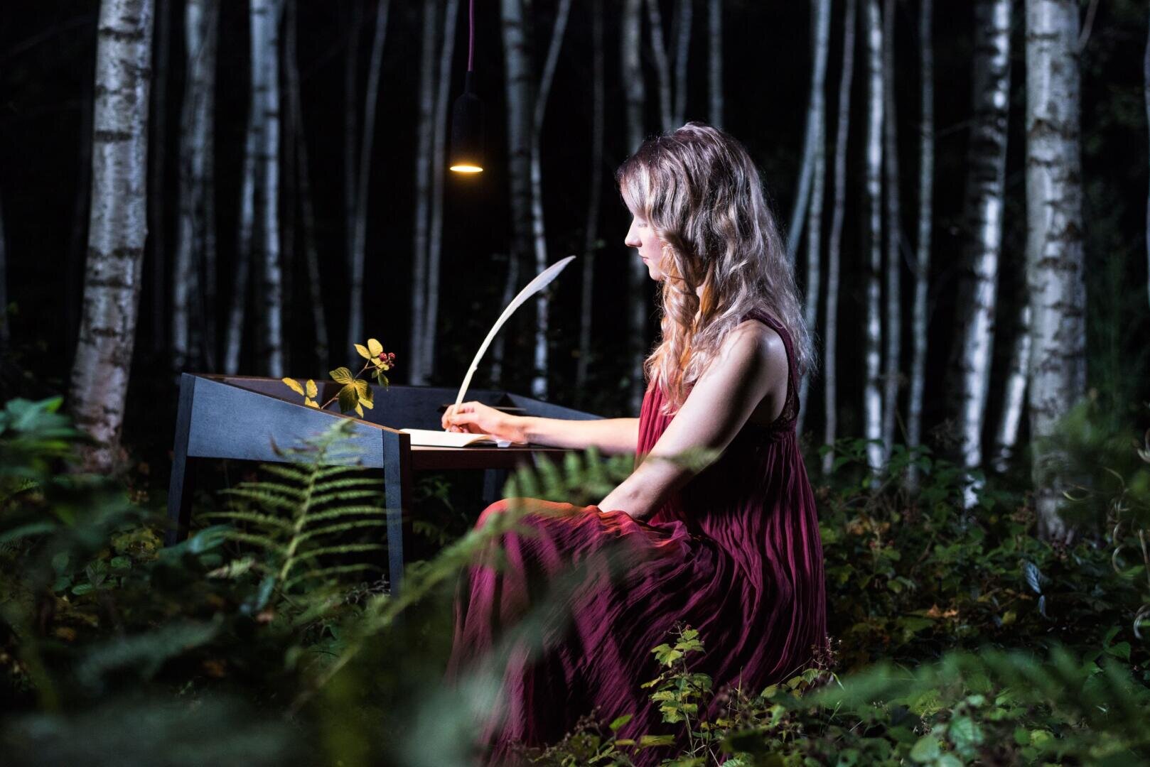 A woman in a flowing burgundy dress sits at a black desk in a forest at night, writing with a quill by a hanging lamp, surrounded by ferns and birch trees.