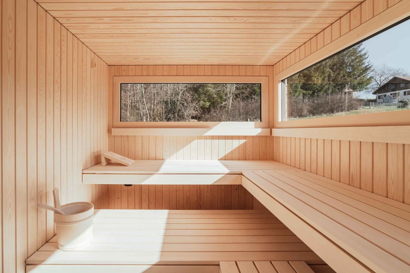 Eine moderne Holzsauna mit großen Fenstern, eingebauten Bänken, einem Holzeimer mit Schöpfkelle und einem ruhigen Ausblick auf Bäume und ein Haus. Das Sonnenlicht strömt durch die Fenster und schafft eine warme und einladende Atmosphäre.