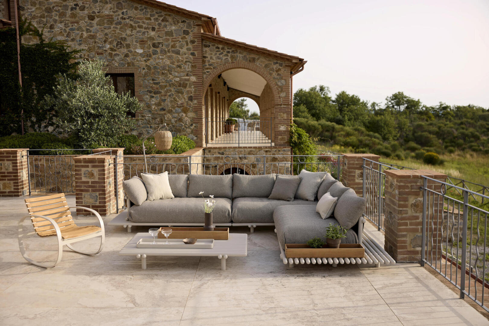 Auf der geräumigen Terrasse mit Stein- und Ziegelakzenten stehen ein modernes Sektionssofa mit grauen Kissen, ein weißer Couchtisch und ein hölzerner Loungesessel mit Blick auf grüne Bäume und ein rustikales Steinhaus.