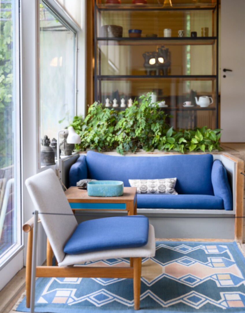 Ein gemütlicher Wohnbereich mit einem blauen Sofa, einem blau-grauen Sessel, einem Holztisch und einem geometrischen blauen Teppich. Hinter dem Sofa stehen grüne Pflanzen, und große Fenster lassen Tageslicht herein. Im Hintergrund sind Regale mit Dekorationen zu sehen.