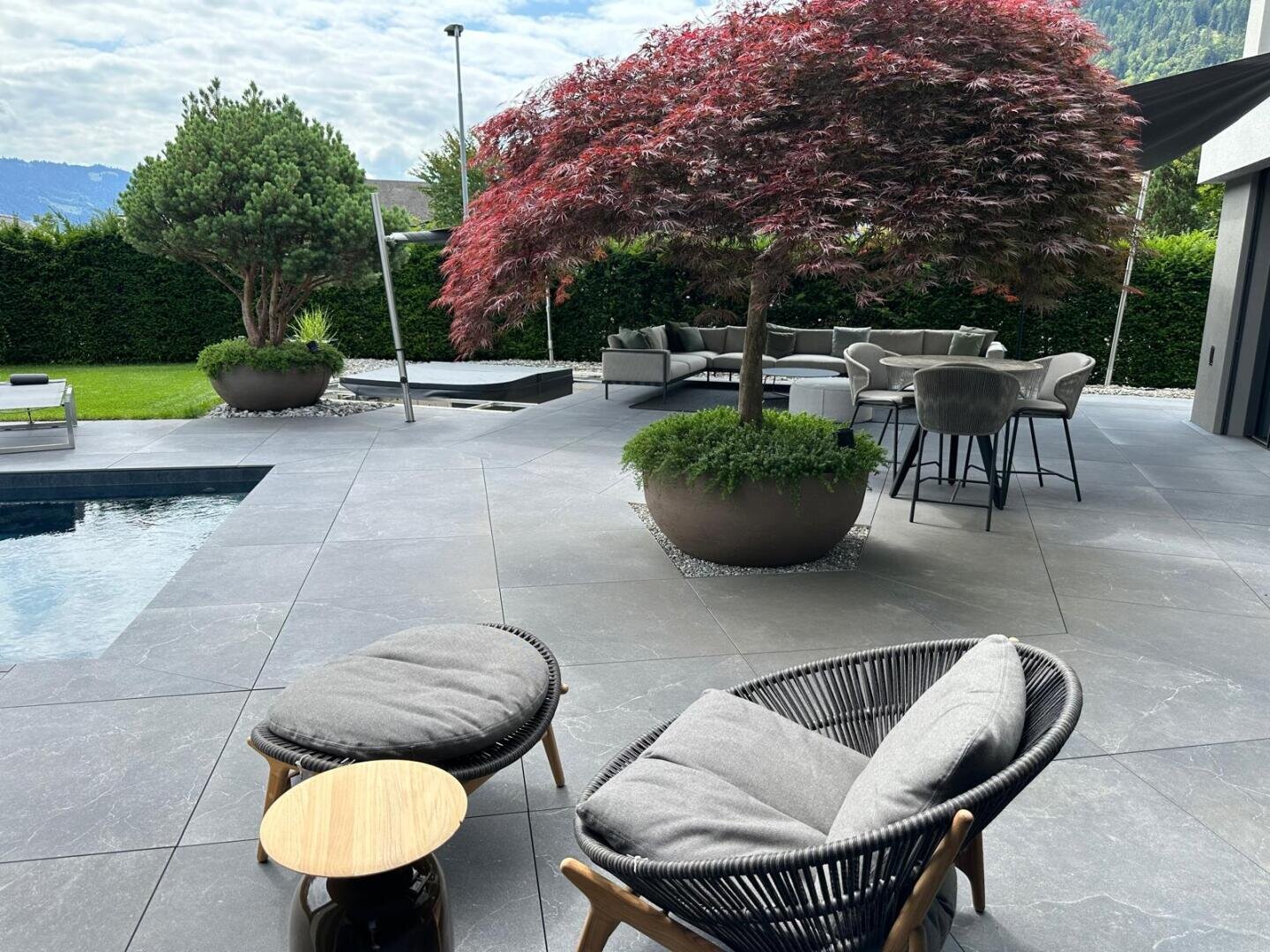 Moderne Außenterrasse mit grauem Fliesenboden, stilvollen Loungesesseln, Topfpflanzen, einem rotblättrigen Baum, einem runden Tisch mit Stühlen und einem Blick auf einen Pool und Grünflächen unter einem teilweise bewölkten Himmel.