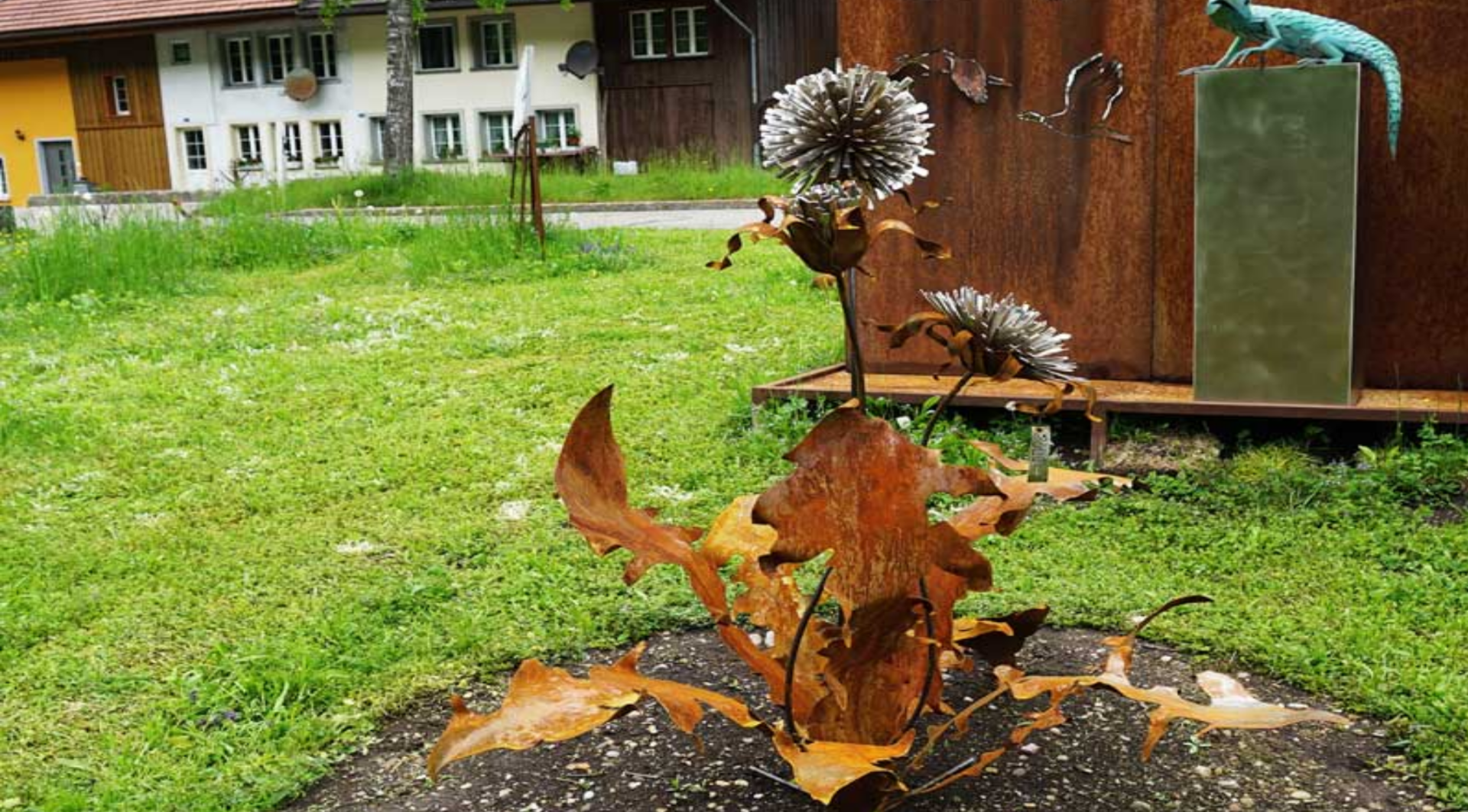 Eine rostfarbene Metallskulptur einer Pflanze mit großen Blättern und runden Blütenköpfen steht im Gras, im Hintergrund sind Gebäude und eine weitere kleine Tierskulptur zu sehen.
