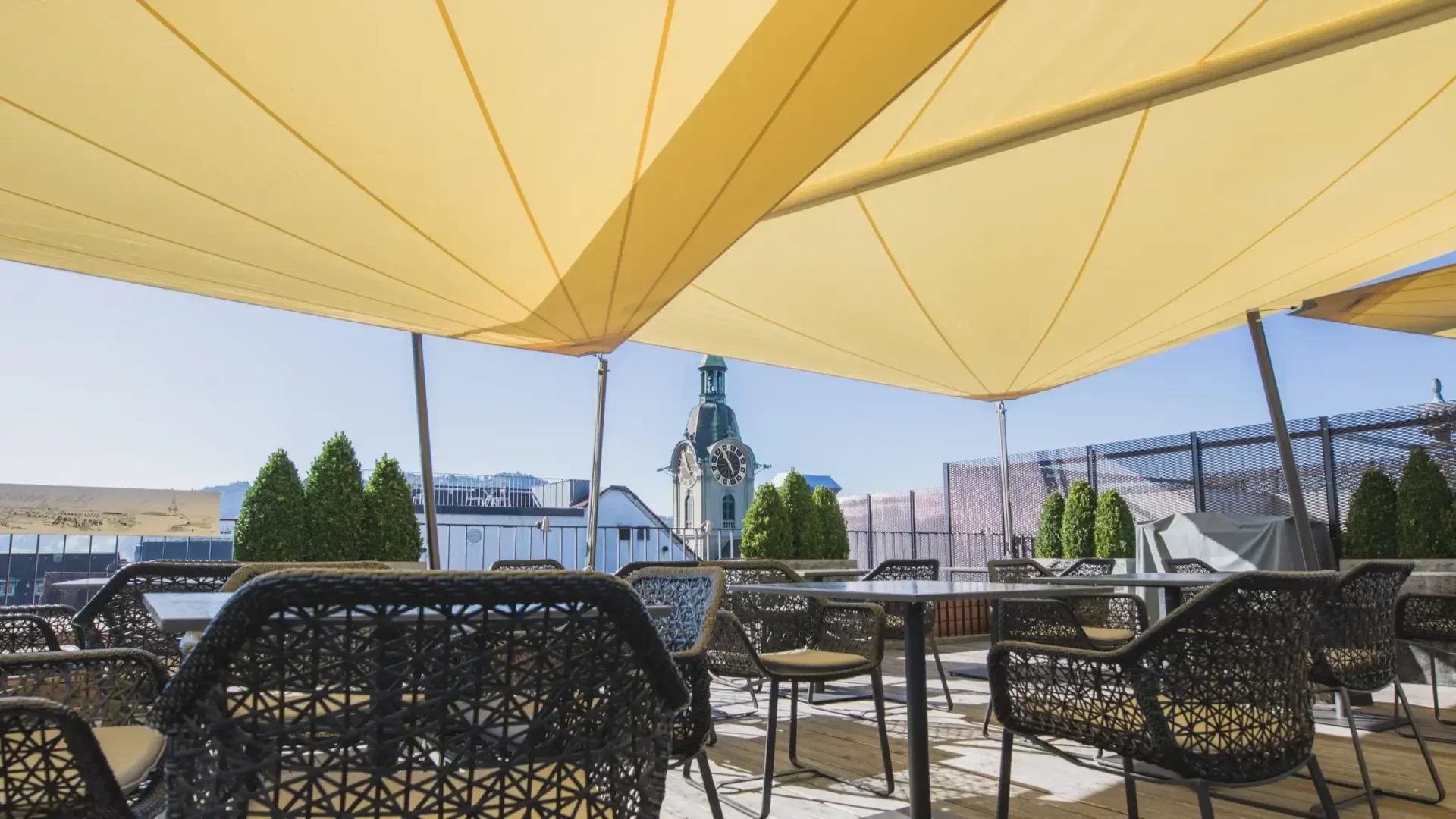 Terrasse mit geflochtenen Stühlen und Tischen unter großen gelben Planen, mit Blick auf die Dächer der Stadt und einen Uhrenturm im Hintergrund, mit klarem blauen Himmel und einigen grünen Sträuchern.