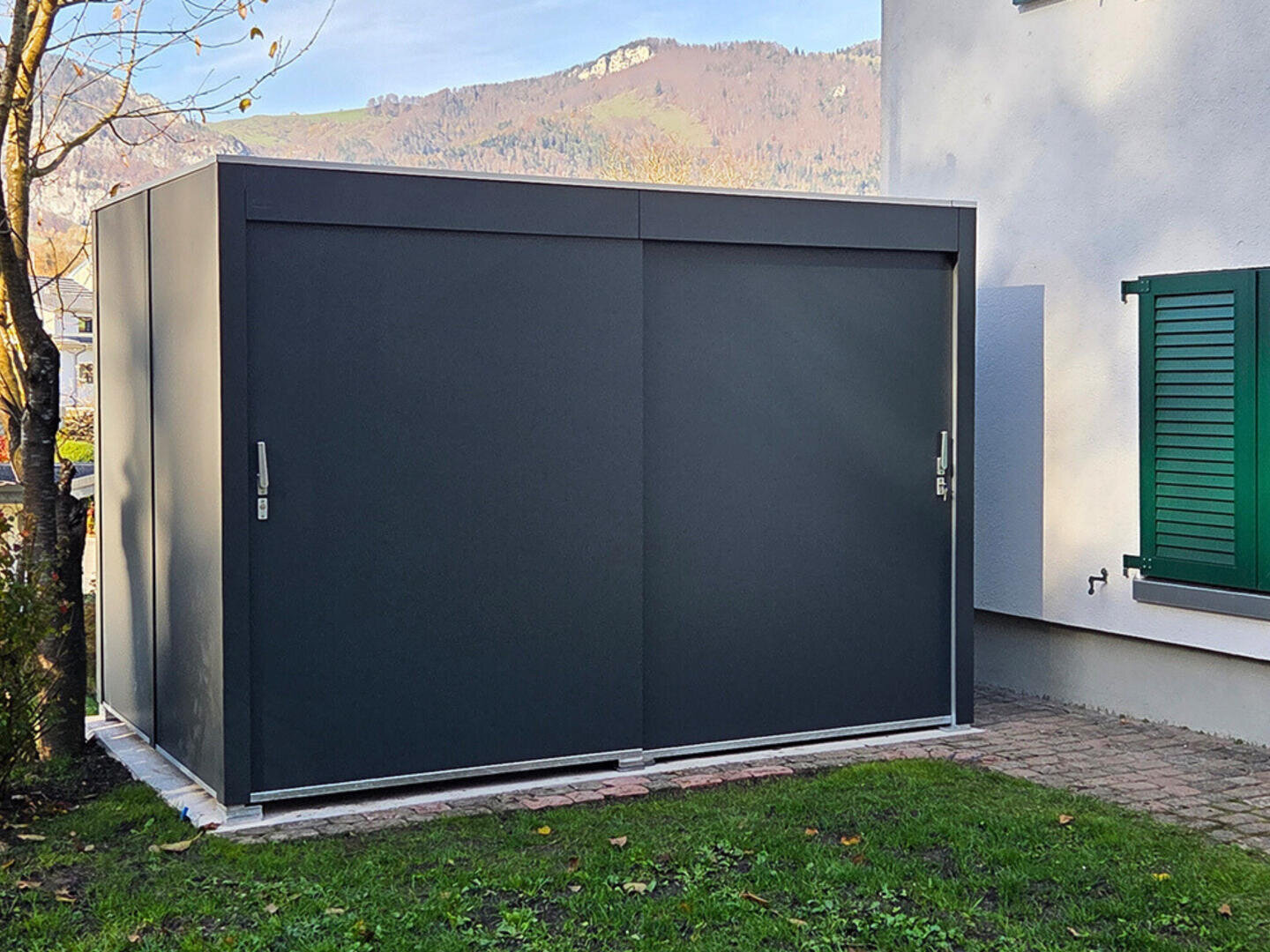 Ein moderner, dunkelgrauer Metallschuppen mit doppelten Schiebetüren steht auf einer gepflasterten Fläche neben einem weißen Haus mit grünen Fensterläden, mit Gras und einem Baum im Vordergrund und Hügeln im Hintergrund.