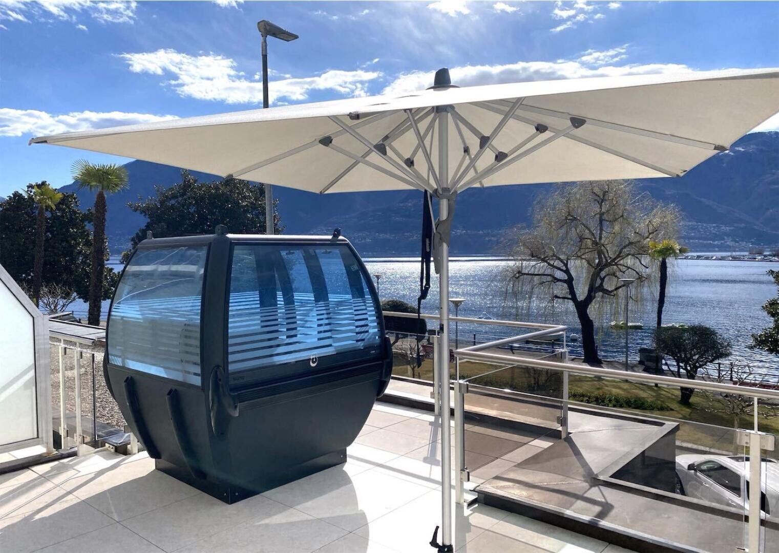 Ein großer weißer Terrassenschirm beschattet eine einzigartige hüttenähnliche Gondel aus Glas und Metall auf einer sonnigen Terrasse mit Blick auf einen malerischen See und die Berge im Hintergrund.