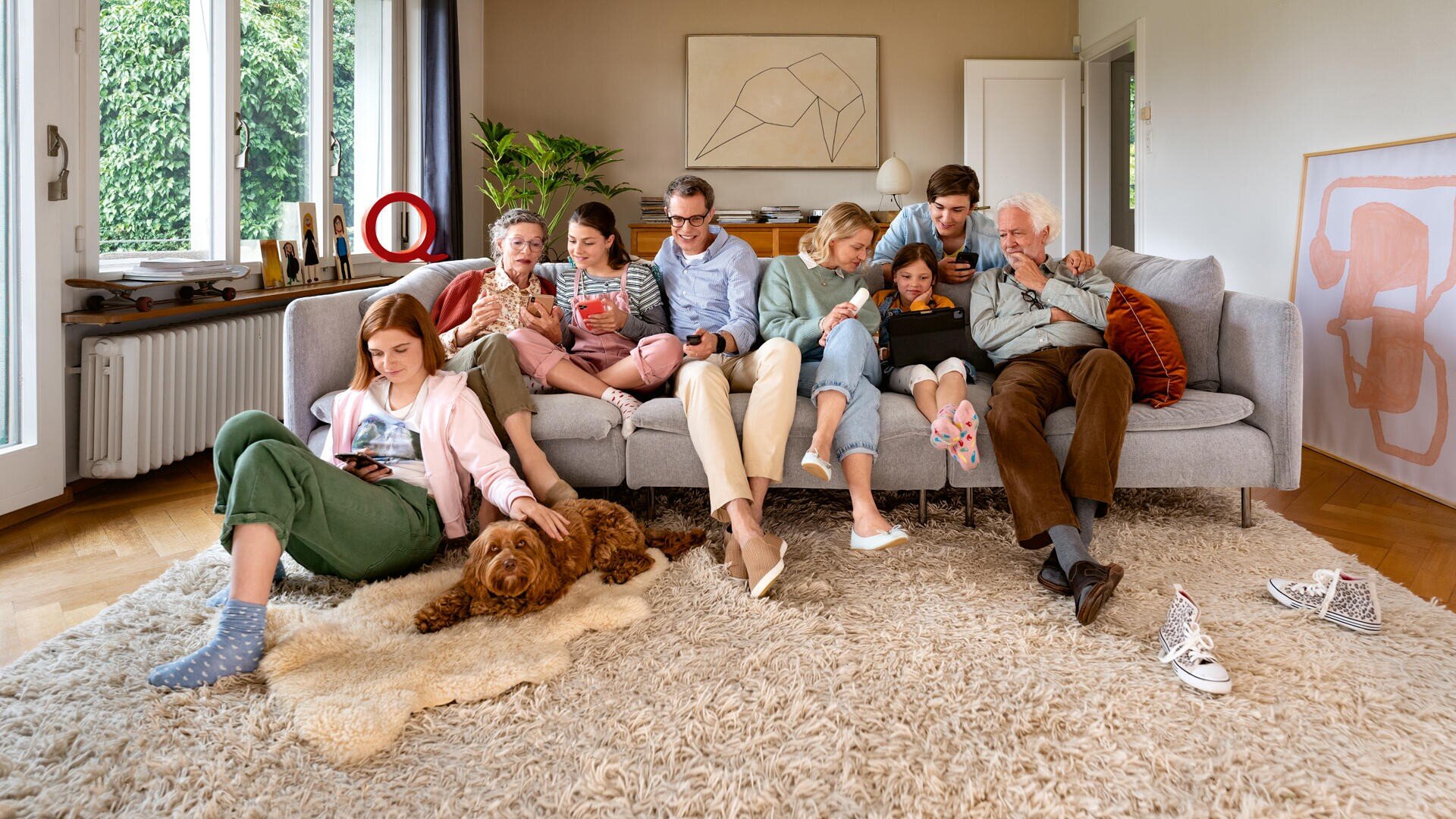 Eine achtköpfige Mehrgenerationenfamilie sitzt eng beieinander auf dem Sofa in einem hellen Wohnzimmer, jeder konzentriert auf sein eigenes Telefon, Tablet oder Laptop. Ein brauner Hund liegt vor ihnen auf dem Teppich.