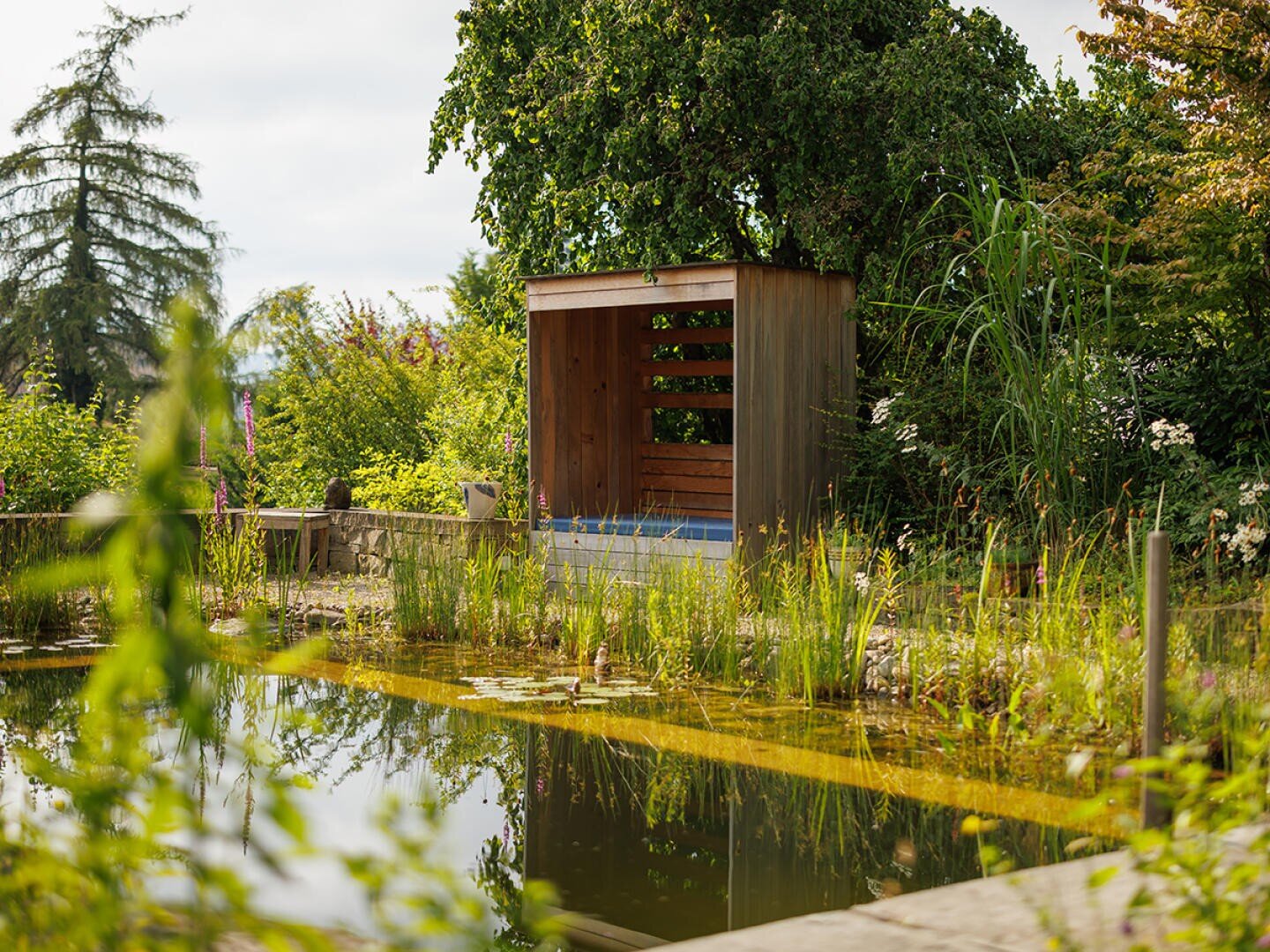 Ein kleiner hölzerner Pavillon steht in der Nähe eines natürlichen Teiches, umgeben von üppigem Grün, hohen Gräsern und Bäumen unter einem teilweise bewölkten Himmel in einer friedlichen Gartenumgebung.
