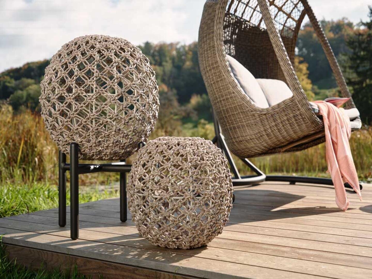 Zwei geflochtene Ottomane stehen auf einer Holzterrasse neben einem Hängesessel aus Korbgeflecht mit Kissen und einer rosafarbenen Decke, im Freien mit Grün und Bäumen im Hintergrund.