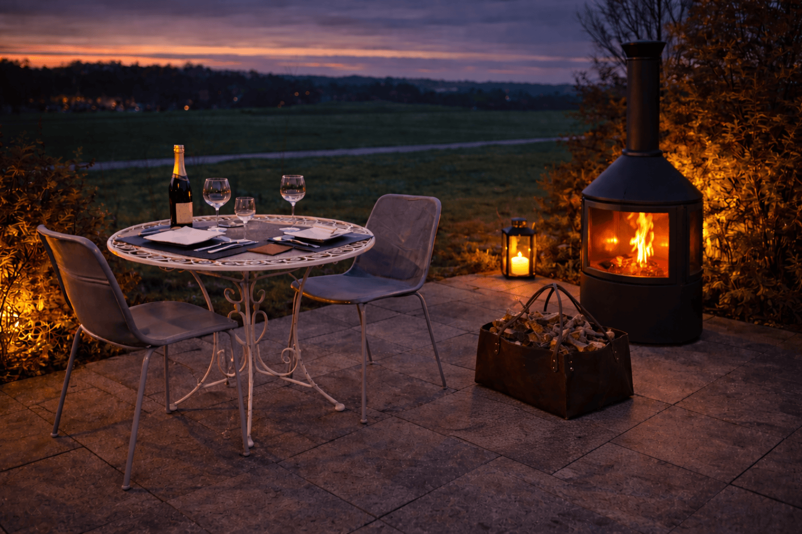 Eine gemütliche Terrassenszene bei Sonnenuntergang mit einem runden Tisch für zwei Personen, Weingläsern, einer Sektflasche und Notizbüchern. Zwei Stühle stehen vor einem beleuchteten Außenkamin mit Laternen und einem Korb mit Brennholz in der Nähe.